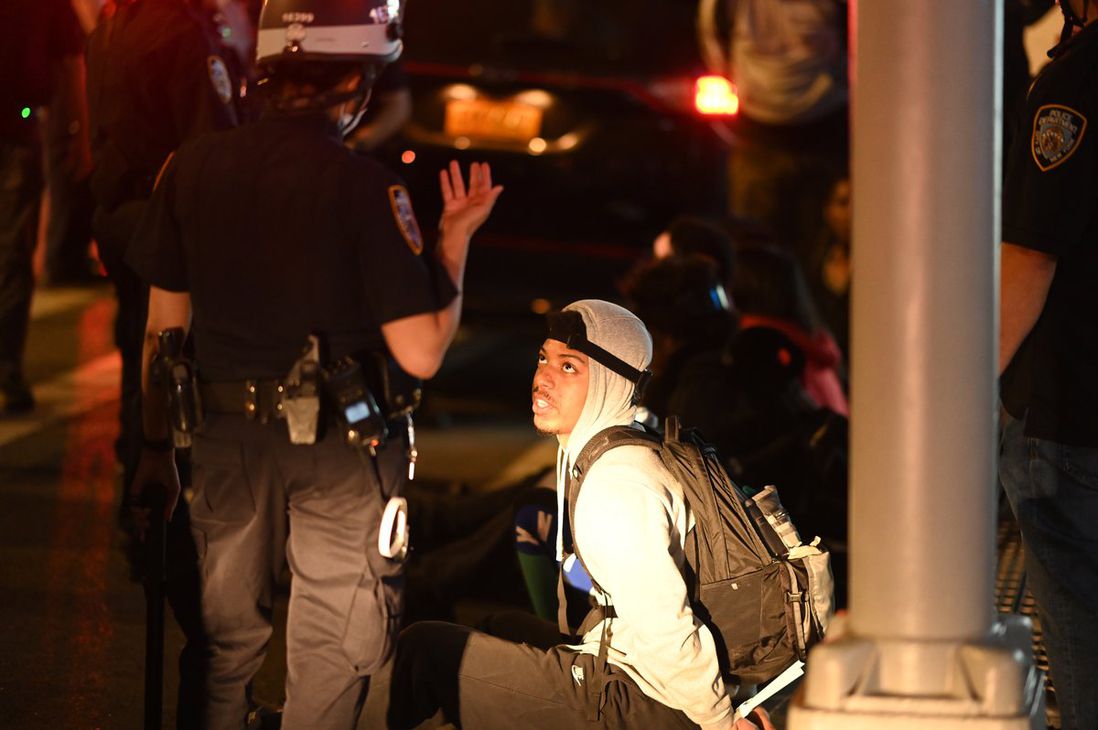 A young black man in handcuffs sitting on the street is surrounded by police.
